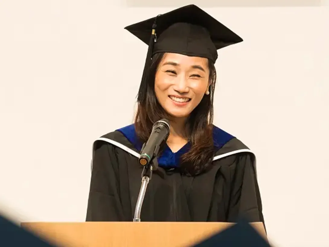 A smiling graduate in a cap and gown delivers a speech at a podium during a business school graduation ceremony.