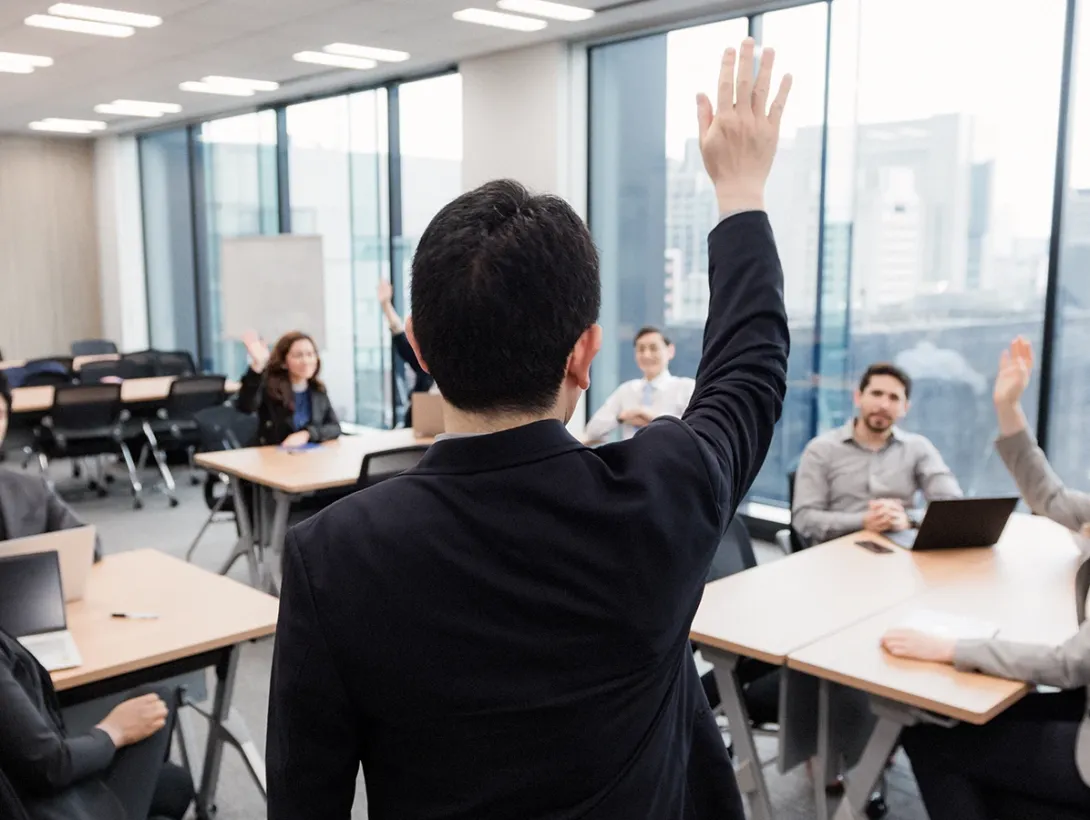 A classroom where a lecturer stands with his hand raised, while several seated students also raise their hands.