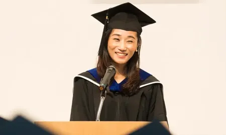 A smiling graduate in a cap and gown delivers a speech at a podium during a business school graduation ceremony.