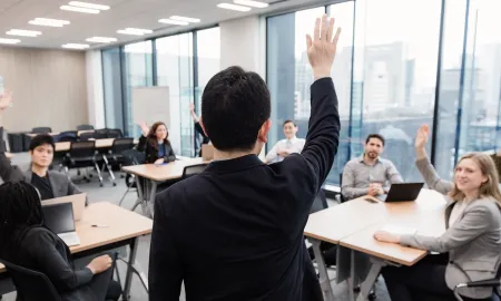 A classroom where a lecturer stands with his hand raised, while several seated students also raise their hands.