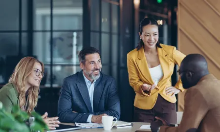 A woman standing leads a diverse group of smiling professionals in a collaborative discussion.