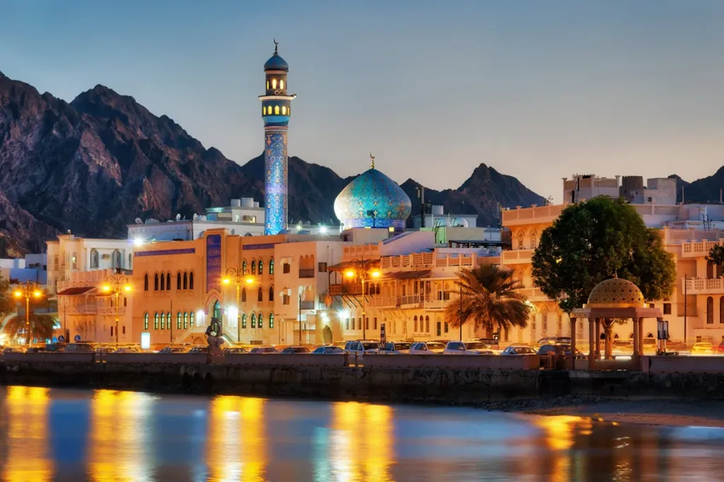 Illuminated waterfront cityscape of Muscat, Oman at twilight, featuring a blue-domed mosque and rugged mountains.
