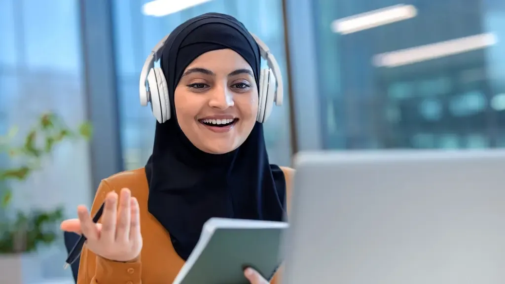 A smiling woman wearing a hijab and headphones gestures while on a video call, holding a notebook and pen.