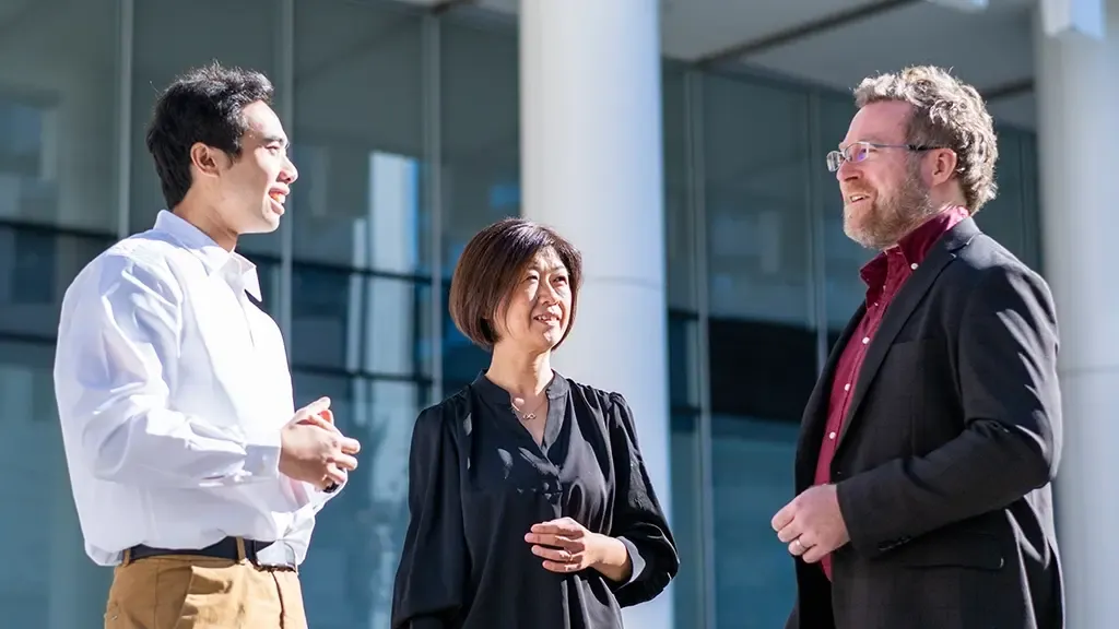 Three diverse students and staff members conversing outside a modern glass business school building.