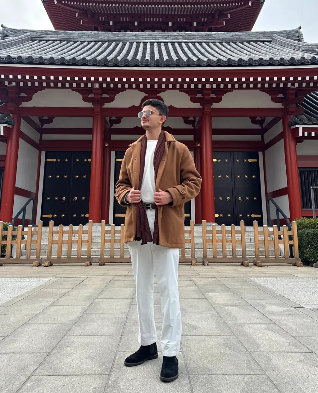 Aiman Alkebsi, wearing a camel coat and burgundy scarf, stands in front of a red-pillared Japanese temple