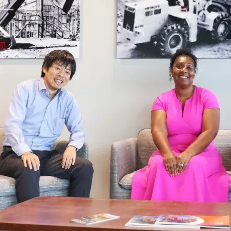 Two smiling professionals seated in a lounge beneath photos of Hitachi machinery.