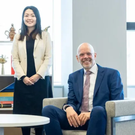 Two professionals pose in a modern office setting; a man is seated in an armchair and a woman stands to his left.
