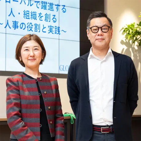 Two smiling professionals standing in front of a presentation screen displaying Japanese text.