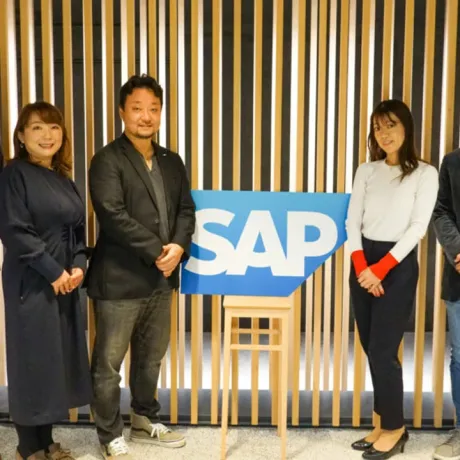 Three professionals standing by a slatted wooden wall, posing next to a large blue and white SAP logo sign.